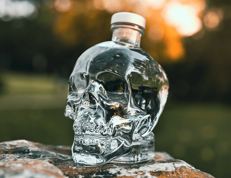 A bottle of Crystal Head Vodka is showcased on a rock with a park blurred in the background